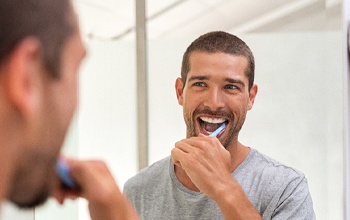 A younger man brushing his teeth while looking at himself in the mirror, happy about his new smile with dental implants