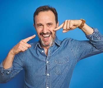 A middle-aged man wearing a blue button-down shirt and pointing to his smile after receiving dentures