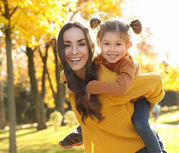 A young woman smiling and showing off her porcelain veneers while giving a little girl a piggyback ride