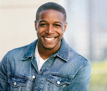 A young man wearing a jean jacket and standing outside while smiling, happy with his veneers 